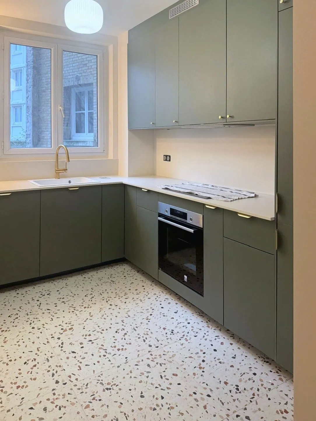 La nouvelle cuisine en L vert sauge vue depuis la porte, plan de travail clair et sol en terrazzo