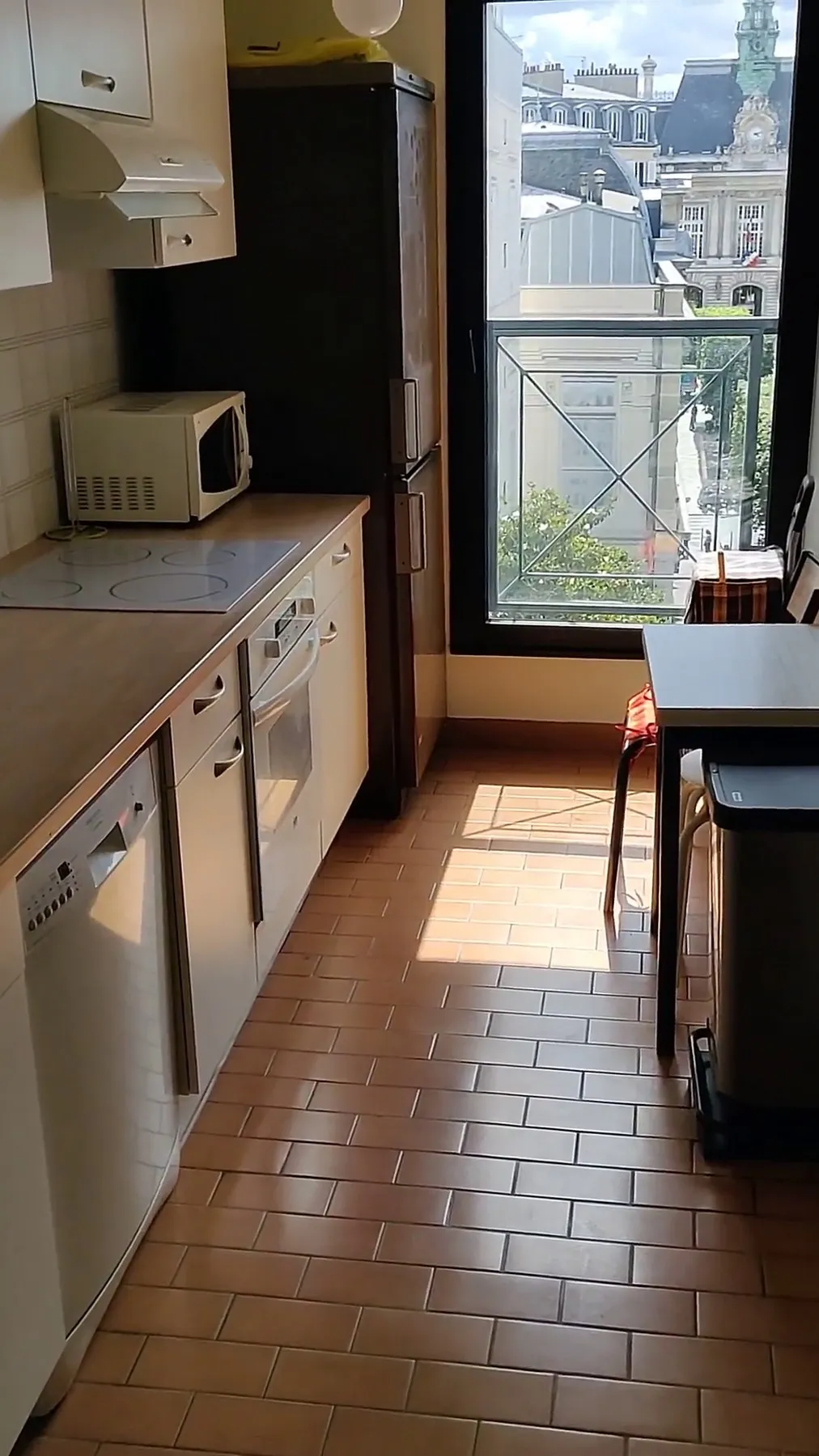 La cuisine avant travaux : pièce en longueur, carrelage brun au sol, meubles alignés jusqu'à la fenêtre