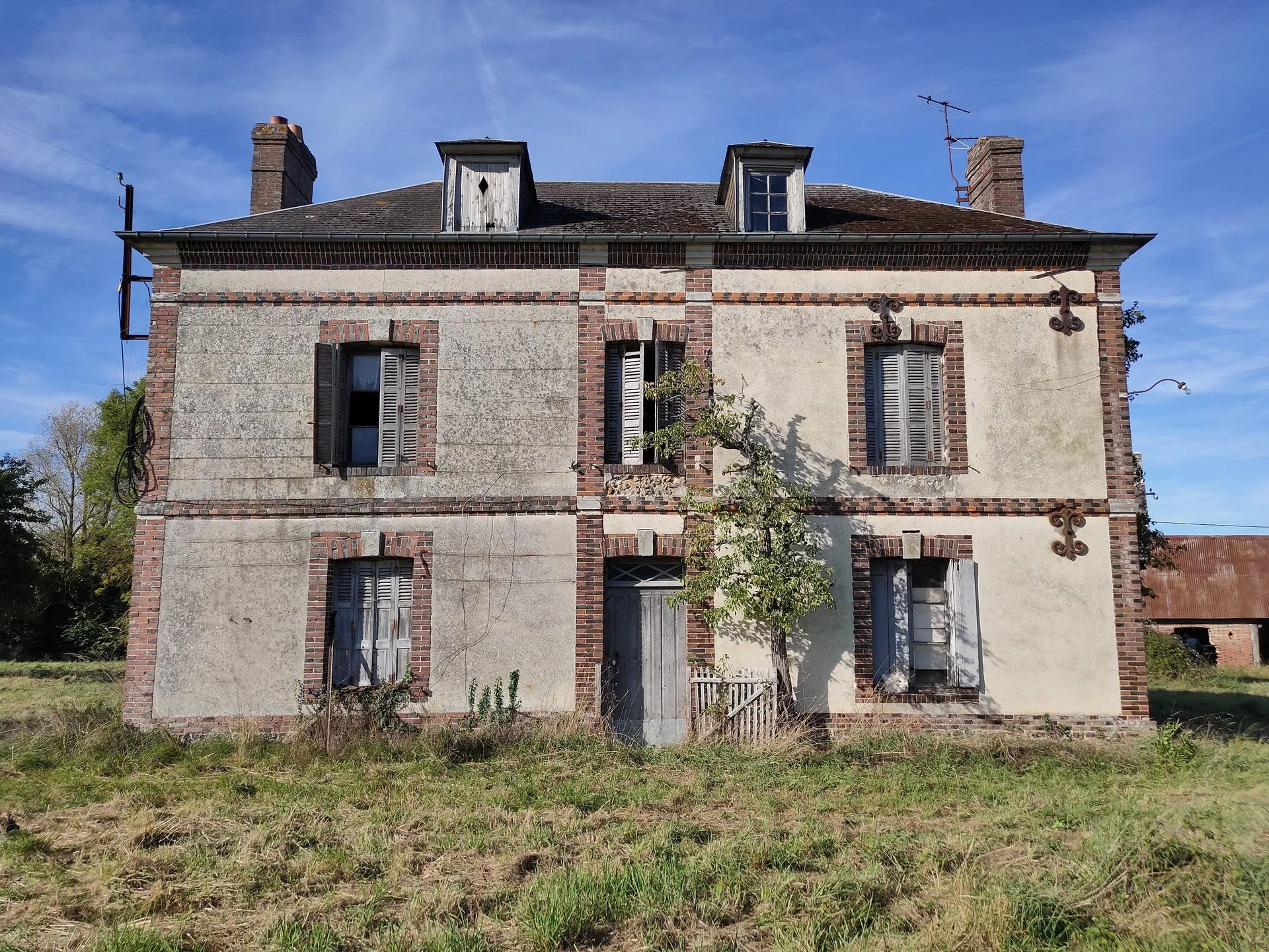 La maison avant travaux, vue du jardin : façade ternie, volets fermés, herbes hautes