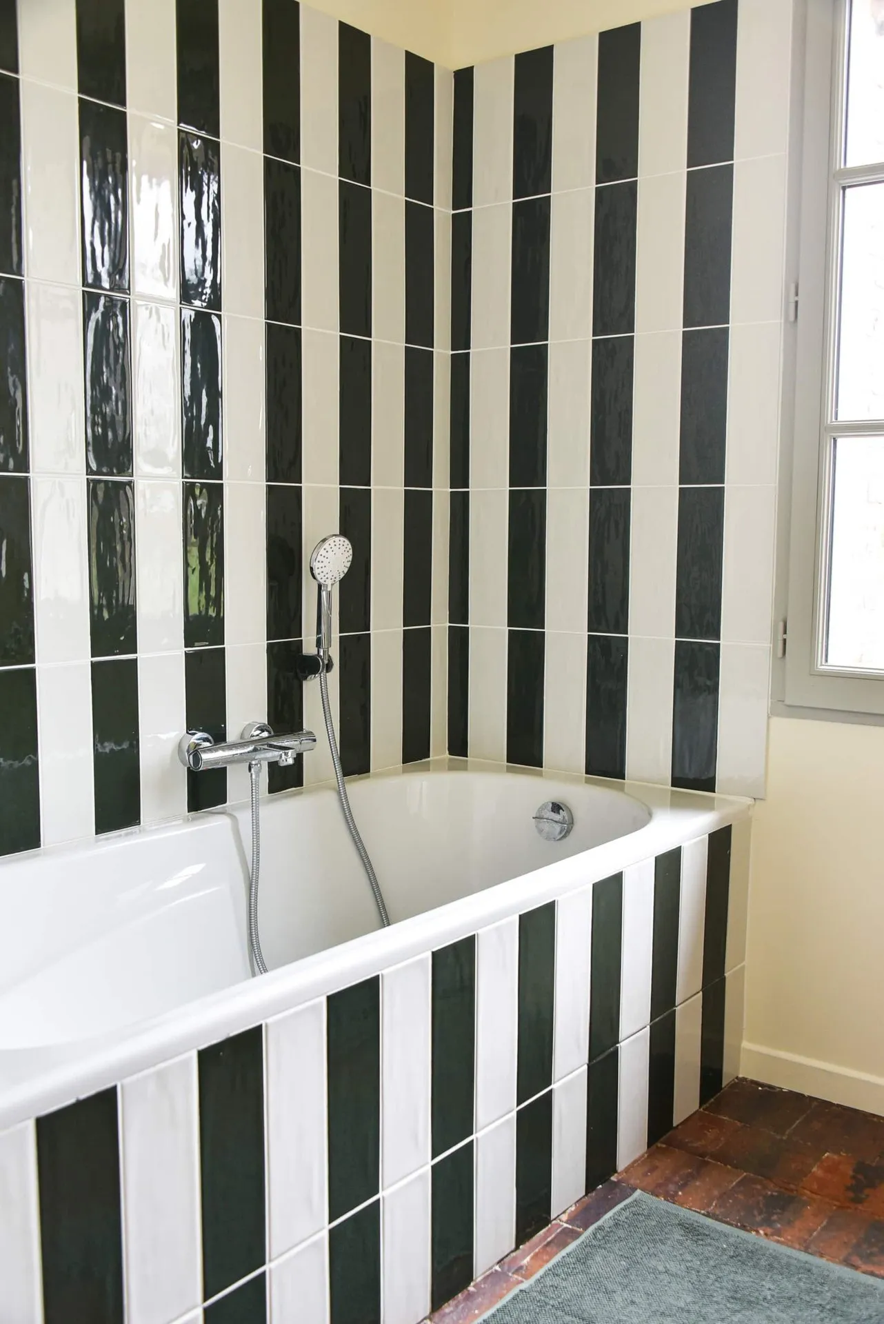 La salle de bain terminée : baignoire et faïence rayée vert sombre et blanc, tomettes au sol